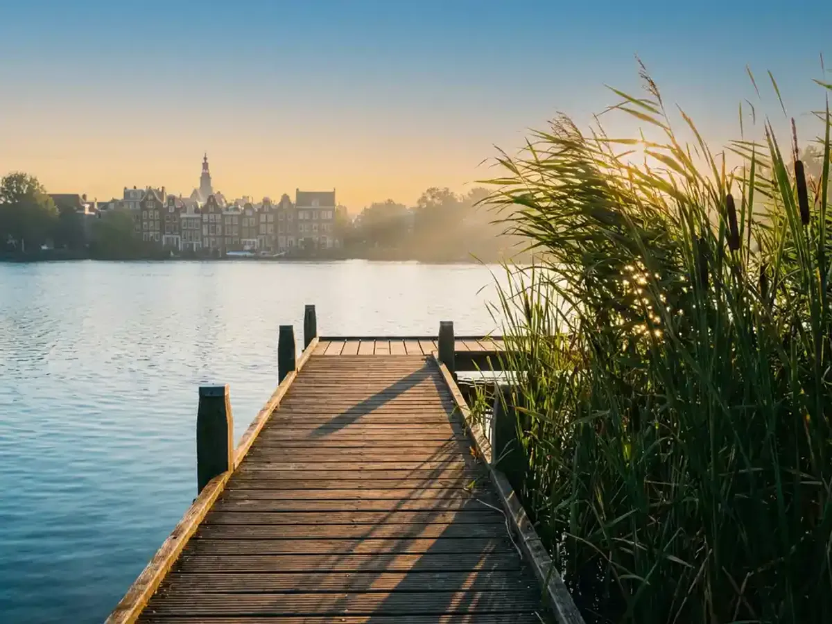 Personeelsfeest in Amsterdam centrum of buiten de stad: voor- en nadelen op een rij? Houten steiger over helder meer met Amsterdamse grachtenpanden in mistige verte, omringd door groene riet