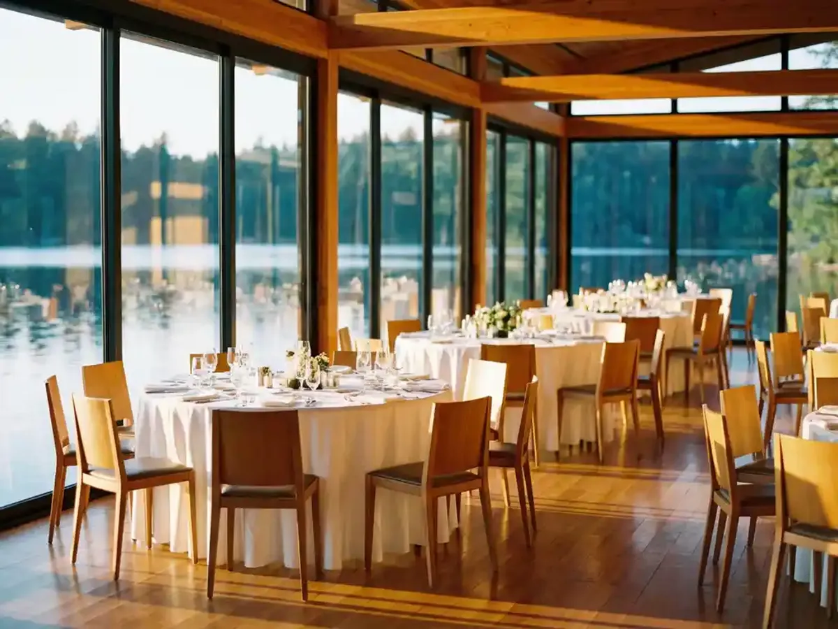 Modern evenementenpaviljoen met glazen wanden en uitzicht op meer, elegante ronde tafels met witte linnen op houten vloer