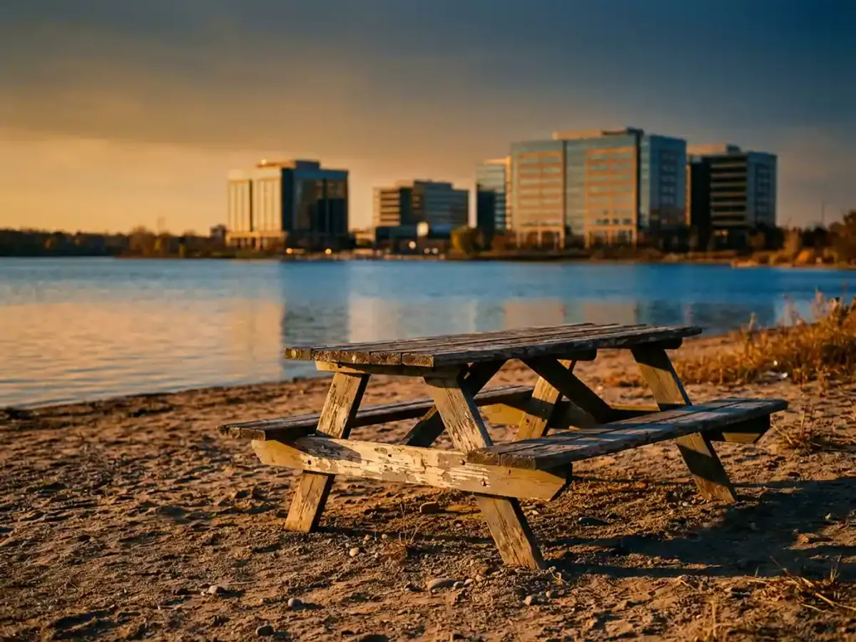 Verweerde houten picknicktafel op zandstrand bij kalm meer met glazen kantoorgebouwen op achtergrond tijdens gouden uur